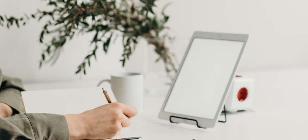 persona en un escritorio con una planta de fondo, escribiendo y con un ordenador en frente