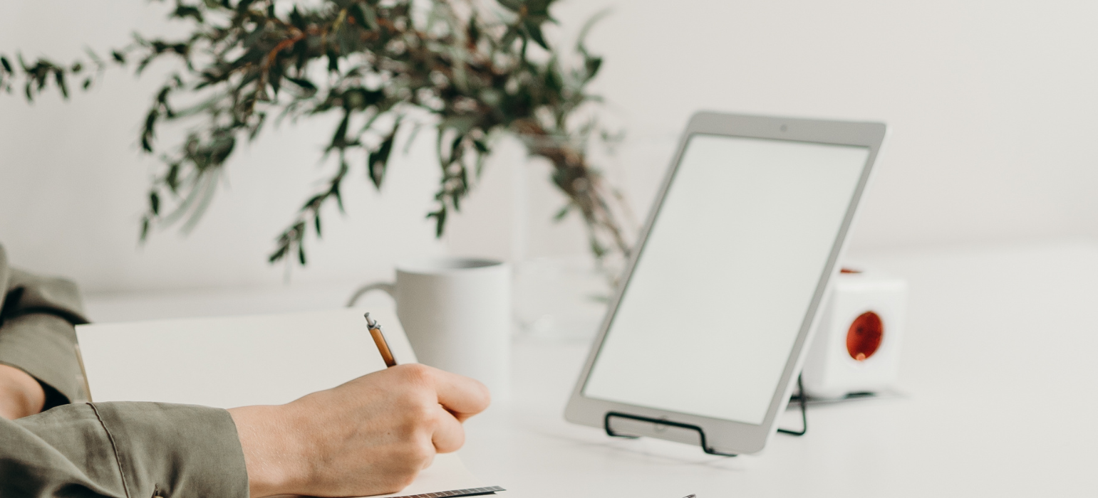 persona en un escritorio con una planta de fondo, escribiendo y con un ordenador en frente